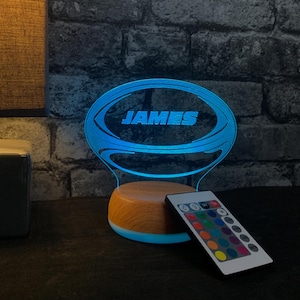 May include: A custom LED night light featuring a blue glowing acrylic rugby ball shape with the name JAMES engraved in the center. The light is mounted on a round wooden base and is accompanied by a multi-colored remote control for lighting.