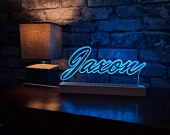 May include: A blue neon sign with the name "Jaxon" in a cursive font. The sign is on a wooden base and is lit up. The sign is in front of a brick wall.