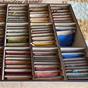 May include: A wooden box with multiple compartments filled with spools of embroidery thread. The box is labeled "Embroidery Threads".