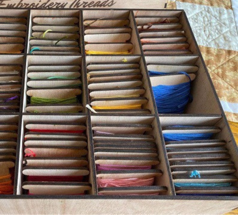 May include: A wooden box with multiple compartments filled with spools of embroidery thread. The box is labeled "Embroidery Threads".
