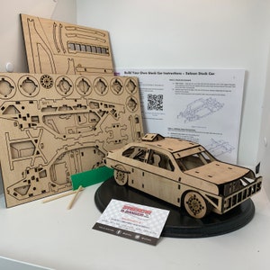 May include: A wooden model kit of a stock car, with the assembled car on a black base. The kit includes instructions and a QR code. The car is made of wood and has a detailed design. The kit is from the company "Stock Car & Banger".