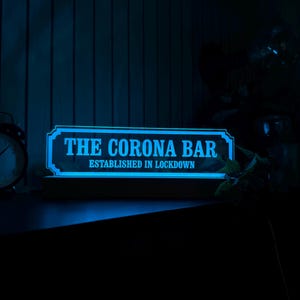 May include: A blue illuminated sign reading "THE CORONA BAR ESTABLISHED IN LOCKDOWN". The sign has a decorative border and is mounted on a wooden base. The background is dark, with a clock and some foliage visible.