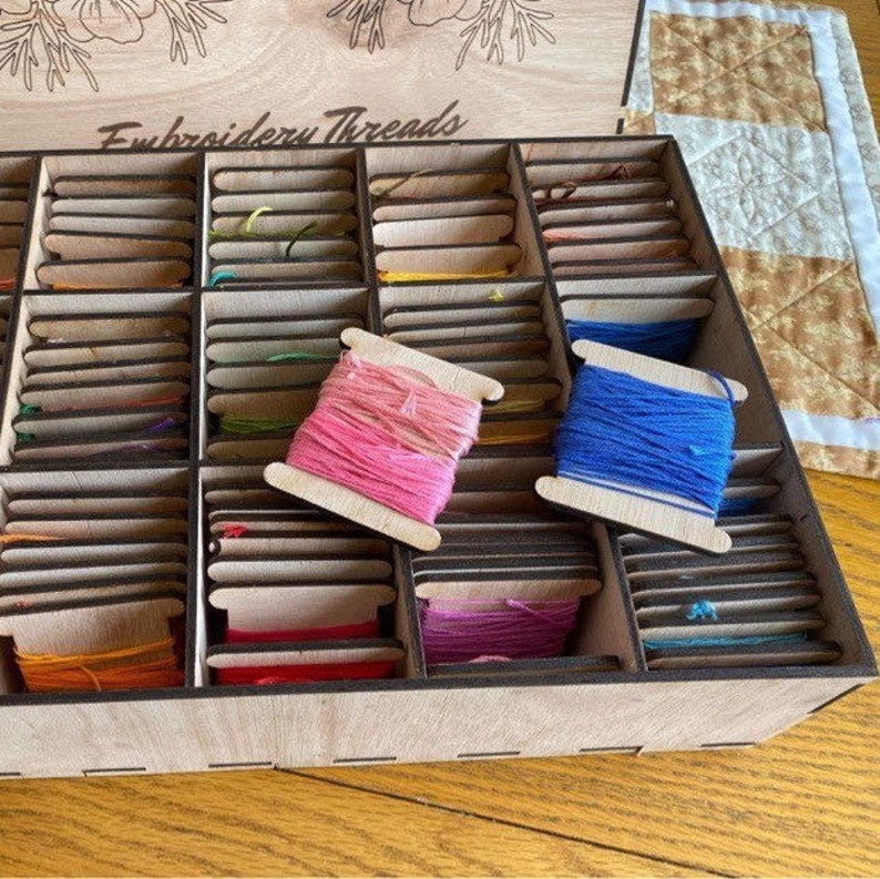 May include: A wooden box with the words "Embroidery Threads" on the lid. The box is filled with small spools of thread in various colors. The spools are arranged in rows and columns.