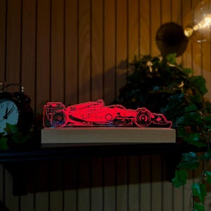 May include: A red LED light display shaped like a Formula 1 race car, featuring the number 16 and sponsor logos on the side. The acrylic silhouette is mounted on a rectangular light-colored wooden base, emitting a vibrant red glow.