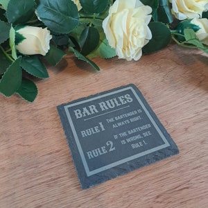 Può includere: Un sottobicchiere in ardesia nera con testo bianco che recita "Bar Rules" e elenca due regole: "Rule 1: The bartender is always right." e "Rule 2: If the bartender is wrong, see rule 1."
