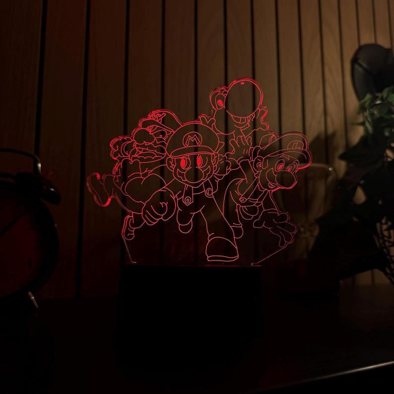 Puede incluir: Una l&aacute;mpara acr&iacute;lica iluminada en rojo con un grupo de personajes de dibujos animados, incluyendo a Mario, Luigi y Yoshi. La l&aacute;mpara est&aacute; sobre una base negra, con un fondo de madera. El dise&ntilde;o es de estilo dibujo lineal.