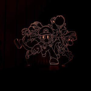 Puede incluir: L&aacute;mpara LED naranja brillante con dise&ntilde;o de Mario, Luigi, Yoshi y Wario. La l&aacute;mpara, con base negra, presenta los personajes en naranja, creando un efecto 3D. Ideal para una sala de juegos o dormitorio infantil.