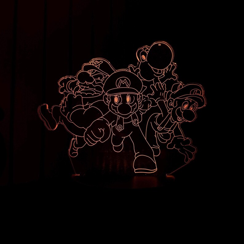 Puede incluir: L&aacute;mpara LED naranja brillante con dise&ntilde;o de Mario, Luigi, Yoshi y Wario. La l&aacute;mpara, con base negra, presenta los personajes en naranja, creando un efecto 3D. Ideal para una sala de juegos o dormitorio infantil.