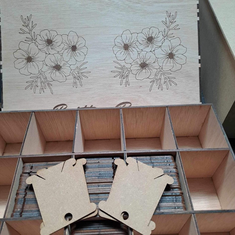 May include: A wooden box with a floral design on the lid. The box has multiple compartments and is filled with small, brown, rectangular pieces of cardboard. The box is perfect for storing small items like thread, beads, or buttons.