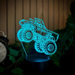 May include: A neon blue monster truck night light with the text "El Toro Loco". The truck has large tires and a bull horn design on top. The light is on a black base and is surrounded by greenery.