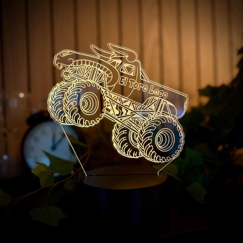 May include: A neon-lit monster truck with the text "El Toro Loco" is displayed. The truck features large, detailed tires and a bull-like design on top. The light is a warm yellow, creating a glowing effect.