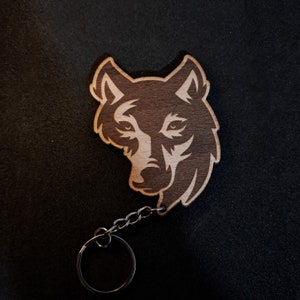 May include: A wooden keychain featuring a laser-cut wolf head design. The wolf is facing forward with its mouth closed. The keychain is attached to a silver metal chain and ring.