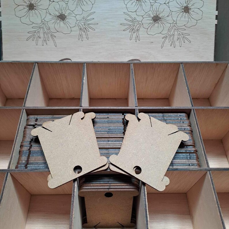 May include: A wooden box with multiple compartments filled with stacks of unfinished wooden thread spools. The spools are light brown and have a hole in the center. The box is made of light wood and has a floral design on the top.