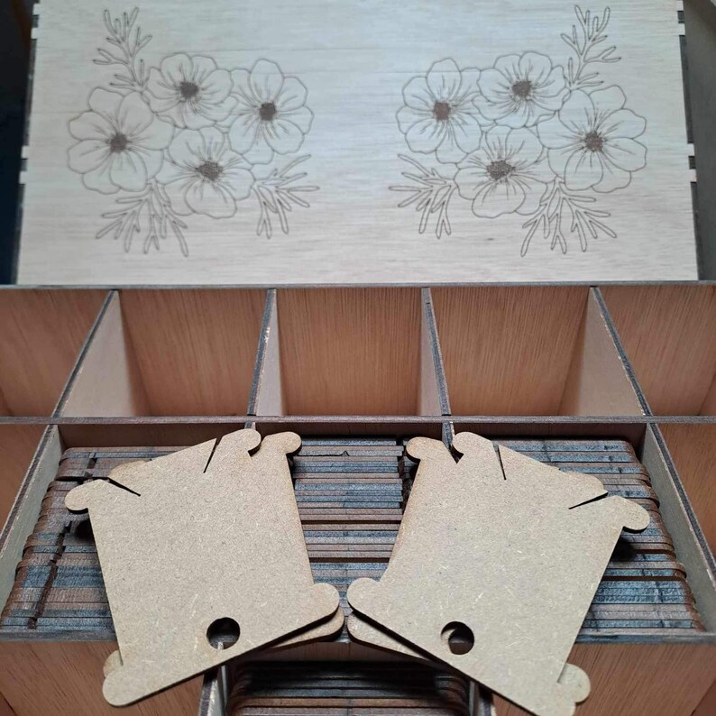 May include: A wooden box with a floral design on the lid. The box has multiple compartments and is filled with small, brown, wooden thread spools.