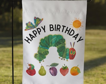 May include: A white garden flag with a green caterpillar, a butterfly, a sun, and the text "Happy Birthday". The caterpillar is eating a variety of fruits and vegetables.