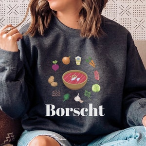 Peut inclure: Une femme portant un sweat-shirt gris foncé avec un graphique d'un bol de bortsch et le mot "Borscht" imprimé sur le devant. Le graphique comprend divers ingrédients comme des betteraves, des oignons, des carottes et de l'aneth.