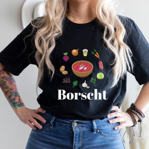 Peut inclure: Un t-shirt noir avec un graphique blanc d'un bol de bortsch entouré d'ingrédients comme des betteraves, des pommes de terre, des carottes et de l'ail. Le texte "Borscht" est imprimé sous le bol.