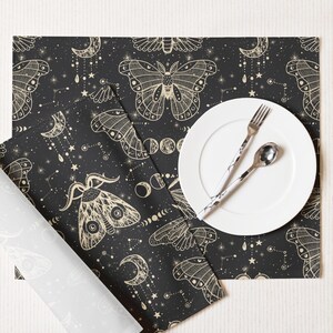 May include: A black placemat with a celestial moth and moon design. A white plate, fork, and spoon are on the placemat. The placemat has a rolled-up napkin with a similar design. The design includes stars and phases of the moon.