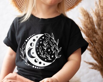 Op de afbeelding: Een zwart T-shirt met een wit halve maan- en sterrenontwerp. De maan is omringd door bladeren en kleine cirkels.