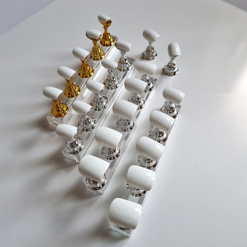 May include: A set of white artificial nails on display stands. The stands are made of clear acrylic and have gold and silver accents. The nails are arranged in rows, with the largest nails at the top and the smallest nails at the bottom.