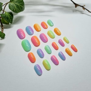 Pode incluir: Uma coleção de unhas ovais de pressão em uma variedade de cores pastel vibrantes. As unhas apresentam um efeito gradiente, passando de uma cor para outra, incluindo tons de rosa, laranja, amarelo, verde e azul. As unhas estão dispostas em uma superfície branca.