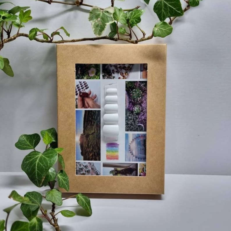 May include: A brown cardboard box with a window showing a set of white press-on nails. The box is surrounded by green leaves.