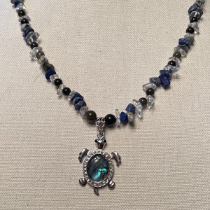 Może przedstawiać: Srebrny naszyjnik z zawieszką w kształcie żółwia z niebiesko-zieloną inkrustacją z abalonu. Naszyjnik wykonany jest z niebieskich, czarnych i przezroczystych koralików z kamieni szlachetnych.