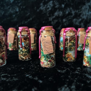 May include: A collection of small glass bottles filled with dried herbs and botanicals. Each bottle has a cork stopper sealed with pink wax and a small tag with the word "away" or "home". The bottles are arranged on a dark, textured surface.