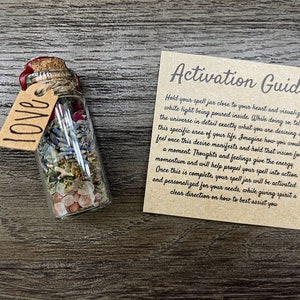 May include: A small glass bottle with a cork stopper and a brown paper tag that says "LOVE" on it. The bottle is filled with dried herbs, flowers, and crystals. A brown card with the text "Activation Guide" is next to the bottle. The card provides instructions on how to activate the spell jar.