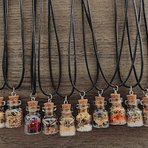 May include: Twelve small glass bottles filled with various dried herbs and flowers, each attached to a black cord necklace. The bottles have cork stoppers and are arranged in a row on a wooden surface.