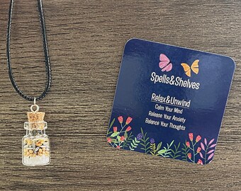 May include: A small glass bottle filled with crystals and beads hangs from a black cord necklace. The bottle has a cork stopper. A blue card with the text "Spells&Shelves Relax&Unwind Calm Your Mind Release Your Anxiety Balance Your Thoughts" is on a wooden surface.