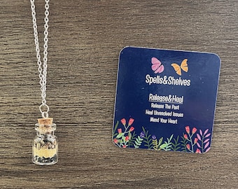 May include: A silver chain necklace with a small glass bottle filled with black and brown crystals. The bottle has a cork stopper. The necklace is on a wooden surface with a blue card that says "Spells&Shelves Release&Heal Release The Past Heal Unresolved Issues Mend Your Heart".