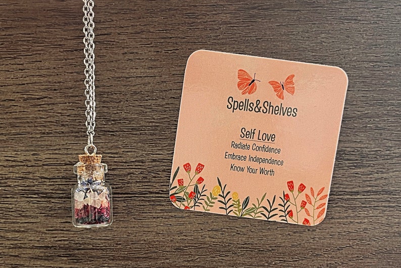 May include: A silver chain necklace with a small glass bottle filled with pink, white, and blue crystals. The bottle has a cork stopper. A card with the text "Spells&Shelves Self Love Radiate Confidence Embrace Independence Know Your Worth" is in the background.