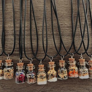 Puede incluir: Doce peque&ntilde;as botellas de vidrio llenas de diversas hierbas y flores secas, cada una unida a un collar de cord&oacute;n negro. Las botellas tienen tapones de corcho y est&aacute;n dispuestas en fila sobre una superficie de madera.