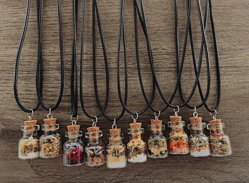 Puede incluir: Doce peque&ntilde;as botellas de vidrio llenas de diversas hierbas y flores secas, cada una unida a un collar de cord&oacute;n negro. Las botellas tienen tapones de corcho y est&aacute;n dispuestas en fila sobre una superficie de madera.