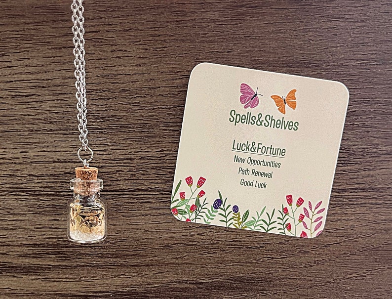May include: A silver chain necklace with a small glass bottle filled with herbs and crystals. The bottle has a cork stopper. The necklace is accompanied by a card that reads "Spells & Shelves Luck & Fortune New Opportunities Path Renewal Good Luck" with a floral border and two butterflies.