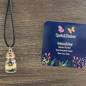 May include: A small glass bottle filled with black and gold crystals hangs from a black cord necklace. The bottle has a cork stopper. A blue card with the text "Spells&Shelves Release&Heal Release The Past Heal Unresolved Issues Mend Your Heart" is in the background.