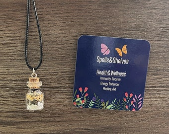 May include: A small glass bottle filled with crystals and herbs on a black cord necklace. The bottle has a cork stopper. The necklace is labeled "Spells & Shelves" and "Health & Wellness: Immunity Booster, Energy Enhancer, Healing Aid."