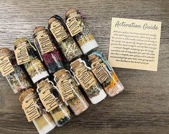 May include: Twelve small glass bottles with cork stoppers and brown paper tags. Each bottle contains a variety of dried herbs, flowers, and crystals. The tags are labeled with words like "Love", "Health", and "Abundance". A small card with the text "Activation Guide" is visible in the background.