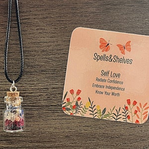 May include: A small glass bottle filled with colourful crystals and dried flowers hangs from a black cord necklace. The bottle is labelled "Self Love" and includes the text "Spells & Shelves".