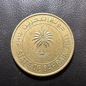 May include: A gold-colored coin with the text "State of Bahrain" and the year "1992" on it. The coin also features the Bahraini coat of arms, which includes a palm tree and a boat.