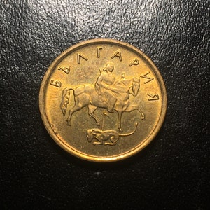 May include: A gold-colored Bulgarian coin with the inscription "БЪЛГАРИЯ" around the edge. The coin features a depiction of a rider on horseback with a dog at the bottom.