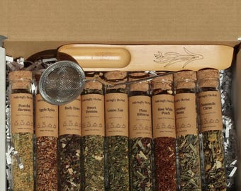 May include: A gift set of herbal teas in glass vials with cork stoppers. The vials are labeled with tea names like "Peaceful Harmony" and "Apple Spice." A wooden tea scoop and a tea infuser are included in the box.