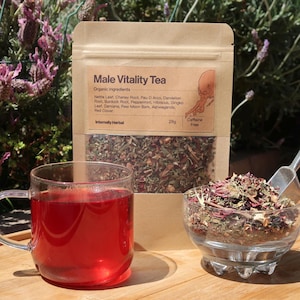 May include: A clear glass mug filled with red tea sits next to a clear glass bowl of loose tea leaves. A brown paper bag labeled "Male Vitality Tea" is behind them. The bag lists organic ingredients and is caffeine-free.