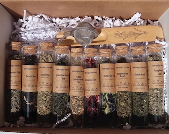 Könnte beinhalten: Geschenkbox mit Glasröhrchen, gefüllt mit verschiedenen Kräutertees, jeweils mit Namen beschriftet. Ein Tee-Ei und ein Holzlöffel liegen obenauf. Die Box ist mit weißen Papierschnipseln gefüllt.
