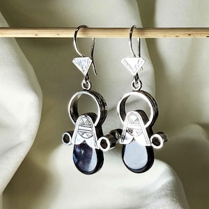 Puede incluir: Un par de pendientes de plata con una parte superior en forma de diamante, una parte media circular y una base en forma de lágrima. Los pendientes tienen un diseño detallado en la sección media y una lágrima oscura y reflectante. Cuelgan de una varilla de madera.