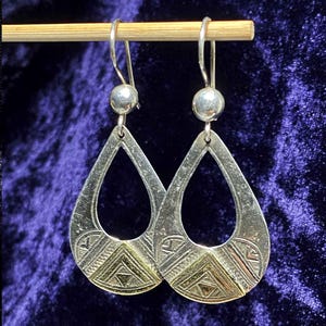 Puede incluir: Pendientes plateados en forma de lágrima con diseño geométrico. Los pendientes tienen una bola plateada en la parte superior y una sección dorada en la inferior. Cuelgan de una clavija de madera sobre un fondo morado oscuro.