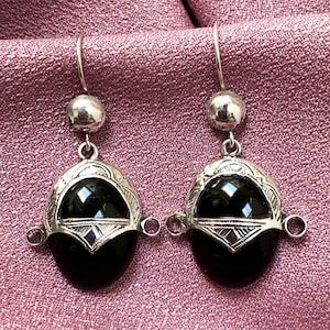 Puede incluir: Un par de pendientes plateados con una piedra negra de forma ovalada. Los pendientes tienen un marco plateado decorativo con patrones intrincados y una pequeña bola plateada en la parte superior. El fondo es una tela rosa texturizada.