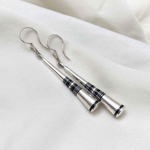 Puede incluir: Un par de pendientes colgantes de plata con incrustaciones negras. Los pendientes son largos y delgados, con una punta puntiaguda.