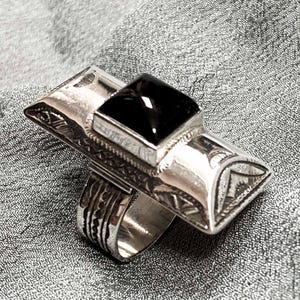May include: A silver ring with a large, rectangular black stone. The ring has an elongated design with etched patterns and a geometric shape around the stone. The ring is set against a gray, textured background.